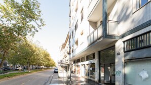 Un immeuble à plusieurs étages, avec un arrêt de bus, des boutiques et une rue bordée d’arbres.