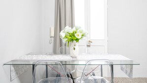Una mesa de vidrio con sillas, un jarrón blanco con flores, una puerta y una ventana con cortinas.