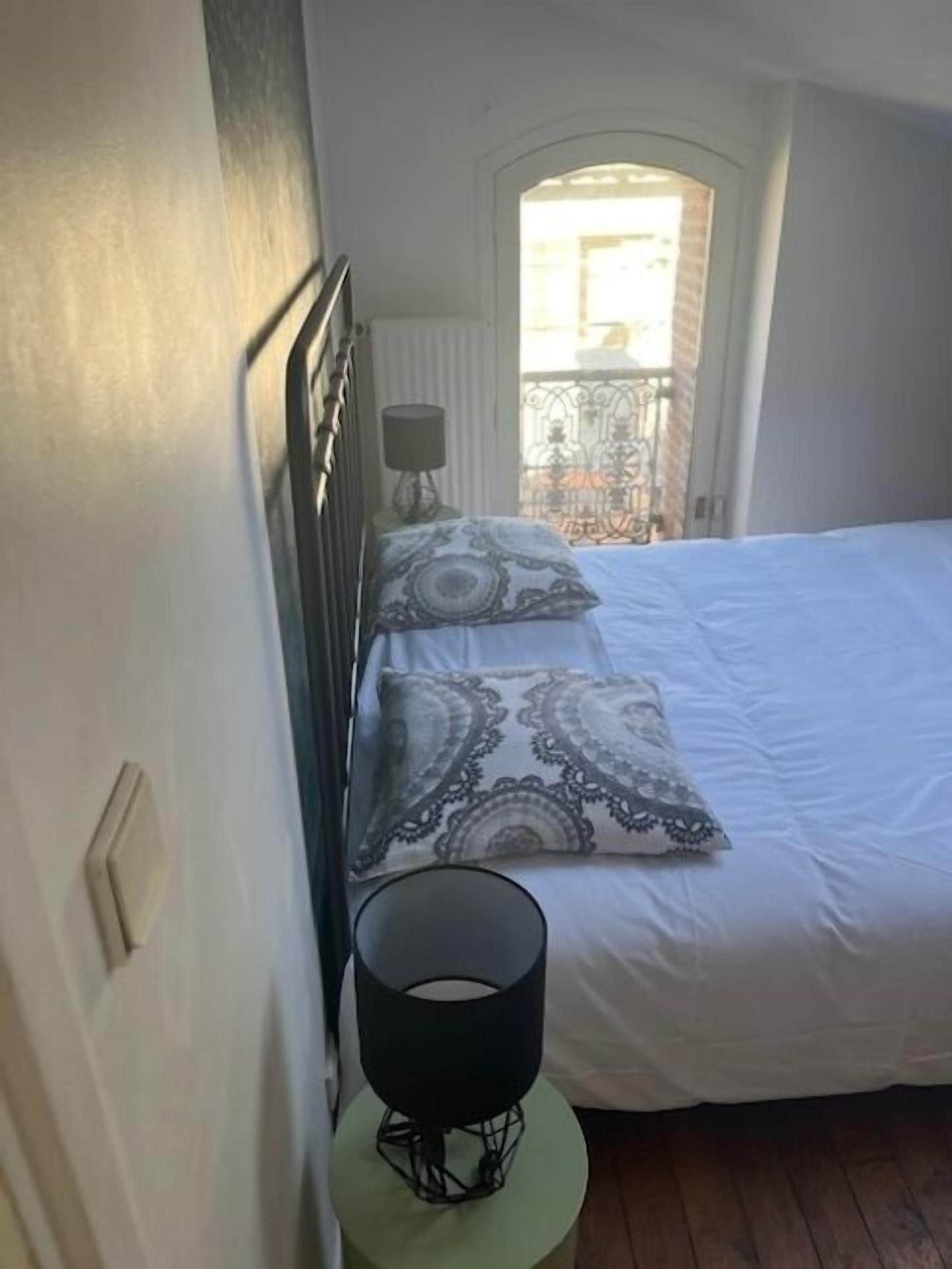 A bedroom with a bed, a bedside table with a lamp, a window, and a radiator.