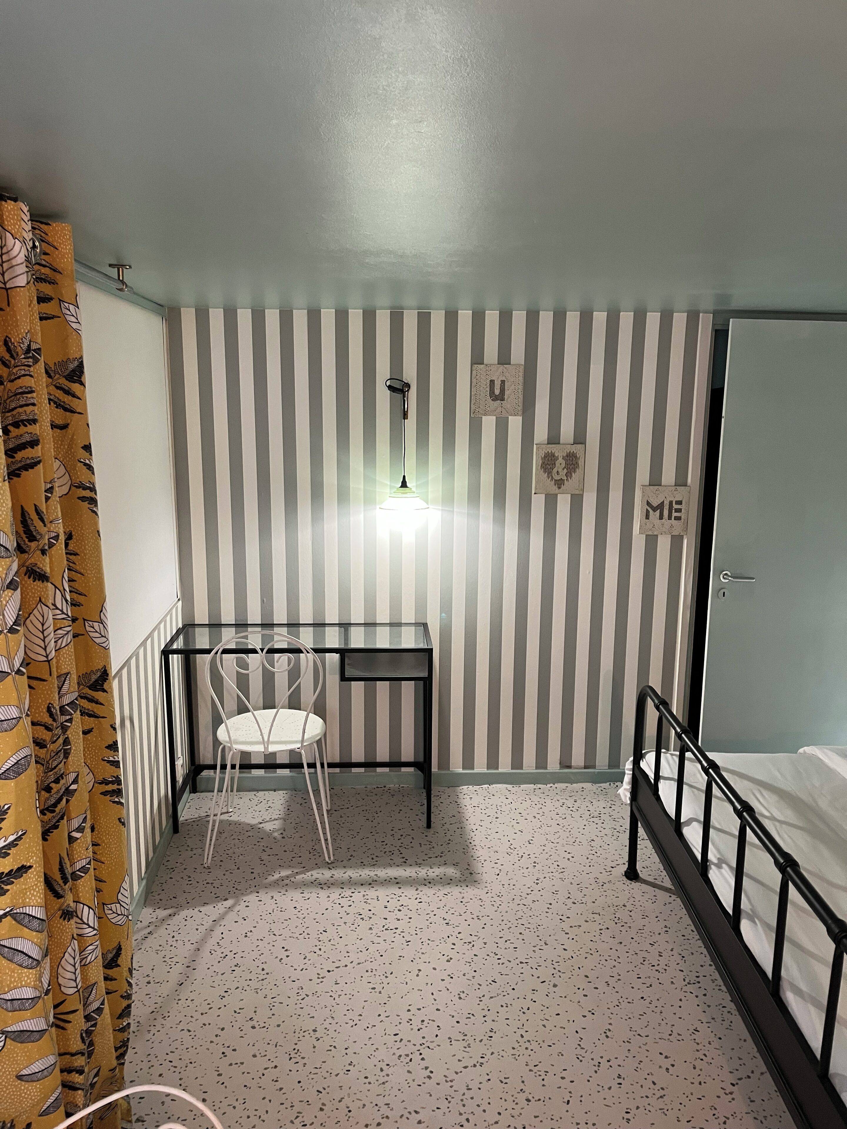 A bedroom with a bed, a desk with a chair, a striped wall, and a curtain.