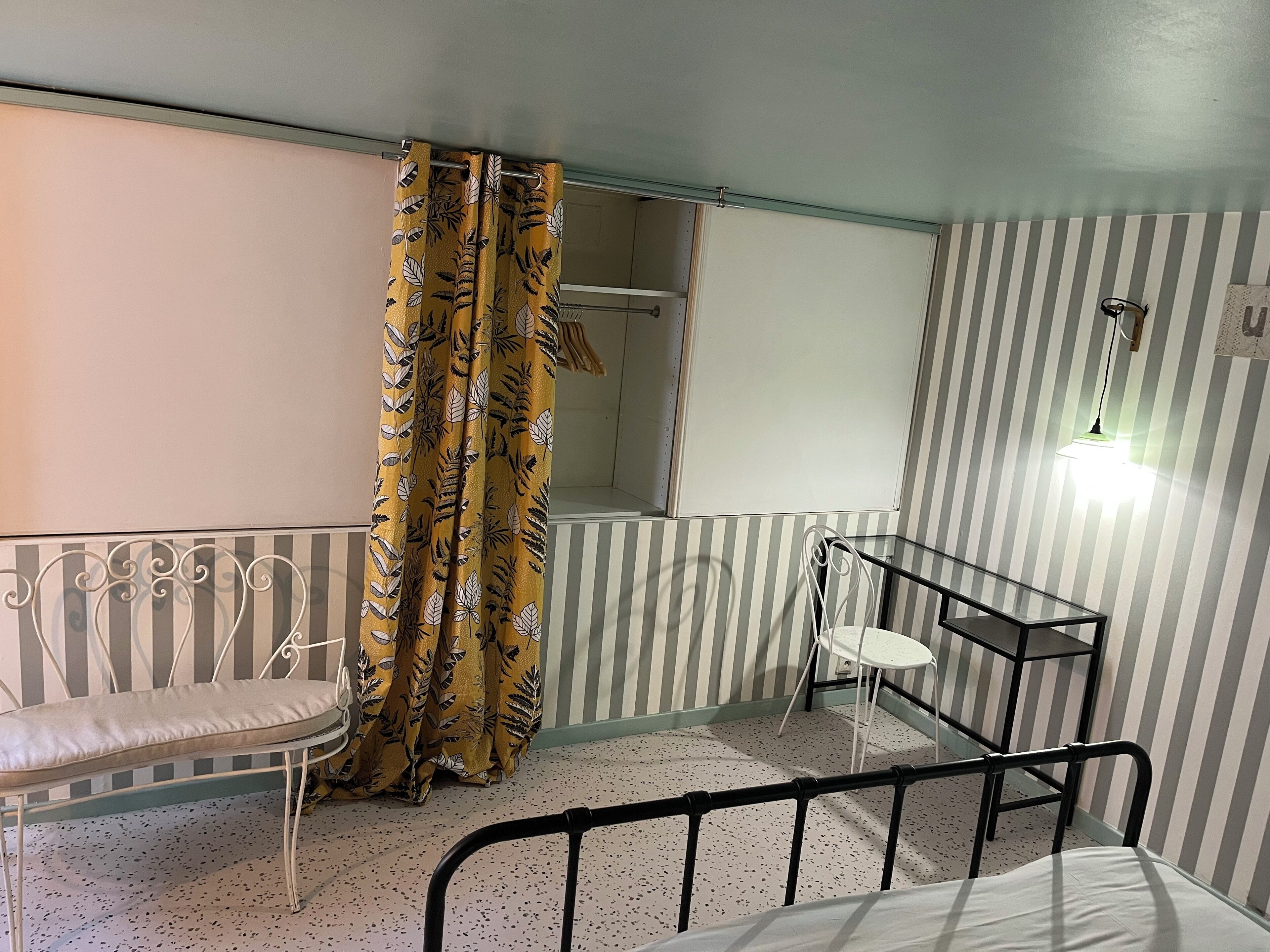 Una habitación con cama, un banco, una silla, pared a rayas, cortina con estampado de hojas y lámpara colgante.