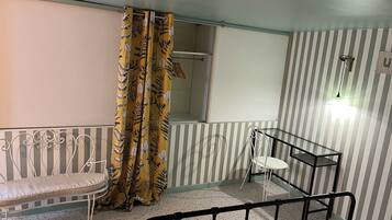 Una habitación con cama, un banco, una silla, pared a rayas, cortina con estampado de hojas y lámpara colgante.