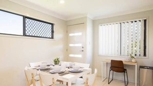 A dining area with a round table set for four, surrounded by white chairs. There is a small side table with a vase and a plant. A window with blinds is visible to the right. - Private Rocklea 2BR | AC | Parking | 220D2 (Rocklea)