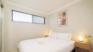A neatly made bed with white linens, a small bedside table with a lamp and a vase of flowers, and a window with blinds. - Private Rocklea 2BR | AC | Parking | 220D2 (Rocklea)
