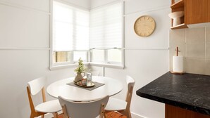 Une cuisine moderne avec des meubles blancs, une table de salle à manger ronde entourée de quatre chaises, une horloge en bois accrochée au mur et un plan de travail noir.