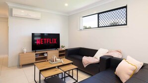 Ein modernes Wohnzimmer mit einem Flachbildfernseher, der 'NETFLIX' zeigt, einer schwarzen Ledersofa, einem Holztisch und einem Fenster mit einem Metalldrahtgitter.