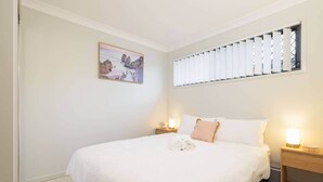 Ein ordentlich bezogenes Bett mit weißer Daunendecke, einem einzelnen rosafarbenen Kissen und einem weißen Handtuch auf dem Boden. An der Wand über dem Bett hängt ein gerahmtes Bild, und zur Rechten befindet sich ein Fenster mit Jalousien.