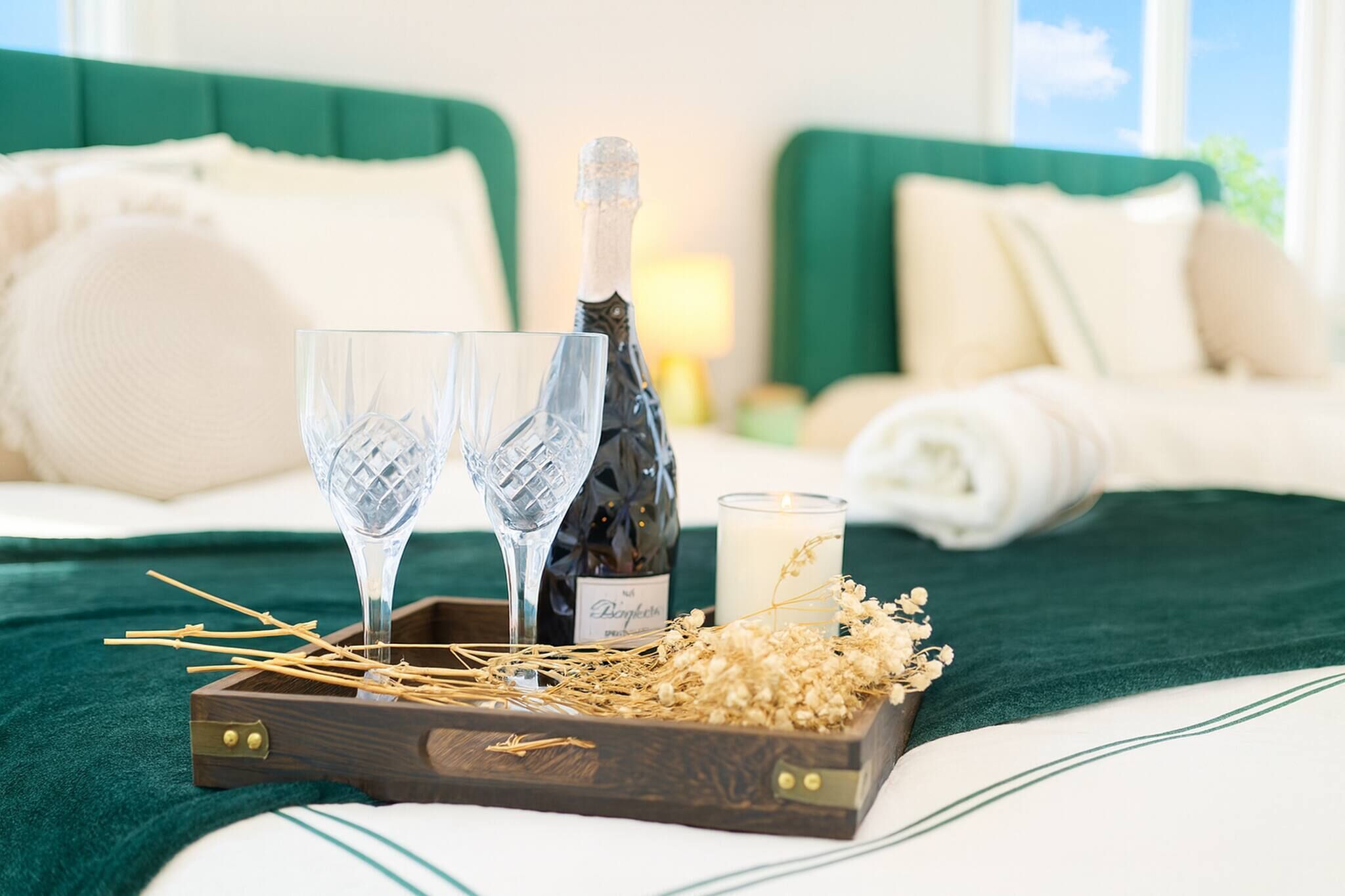 A bed with a green headboard, a wooden tray with a bottle of champagne, two glasses, and a candle.
