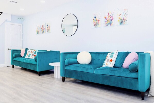 Un canapé de couleur turquoise agrémenté de coussins à motifs floraux, un miroir rond et des œuvres d’art murales abstraites représentant des fleurs, le tout dans une pièce lumineuse au sol en bois clair.