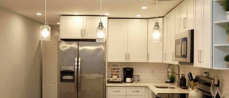 A modern kitchen with white cabinets, stainless steel appliances, pendant lighting, and a coffee station.