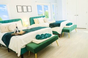 Une chambre avec deux lits, chacun doté d’une tête de lit recouverte d’un tissu vert et de linge de lit blanc. On peut voir des tableaux encadrés au mur, une fenêtre donnant sur des arbres et une porte au fond de la pièce.