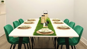Una mesa de comedor con sillas verdes, un mantel blanco y un centro de mesa con una cesta tejida y una planta pequeña.