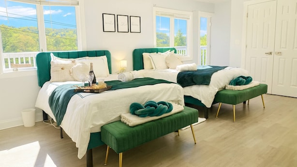 Un dormitorio con dos camas, cabeceros tapizados en verde, ropa de cama blanca y vistas a zonas verdes a través de amplias ventanas.