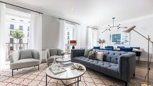 A modern living room with a grey sofa, a round coffee table, and a blue armchair. There is a large window with curtains, a floor lamp, and a framed picture on the wall. - Alcala IX - Three Bedroom Apartment, Sleeps 6 (Madrid)