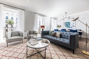 Ein modernes Wohnzimmer mit grauem Sofa, einem runden Couchtisch und einem blauen Sessel. Ein großes Fenster mit Vorhängen, eine Stehlampe und ein gerahmtes Bild an der Wand sind ebenfalls zu sehen.