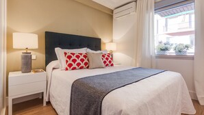A hotel room with a bed, two bedside tables, a nightstand, an air conditioning unit, and a window with a view of a balcony. - General Pardiñas III (Madrid)