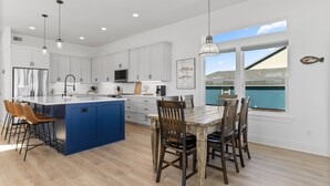 A modern kitchen with white cabinets, a blue island, and a dining area with wooden chairs. - Modern Matagorda Getaway – 4 King Bedrooms, Canal Access & Minutes to the Beach (Matagorda)