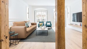 Ein modernes Wohnzimmer mit einem beige gefärbten Sofa, zwei smaragdgrünen Sesseln, einem Couchtisch und einem Fernseher auf einem Holzregal.