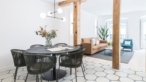 Ein modernes Wohnzimmer mit einem runden Esstisch, schwarzen Korbstühlen, einem braunen Sofa und einem blauen Sessel.