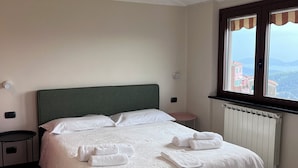 A neatly made bed with white linens and towels, a green headboard, a bedside table with a lamp, and a window with a view of buildings and mountains. - Collimare rooms (Vezzano Ligure)