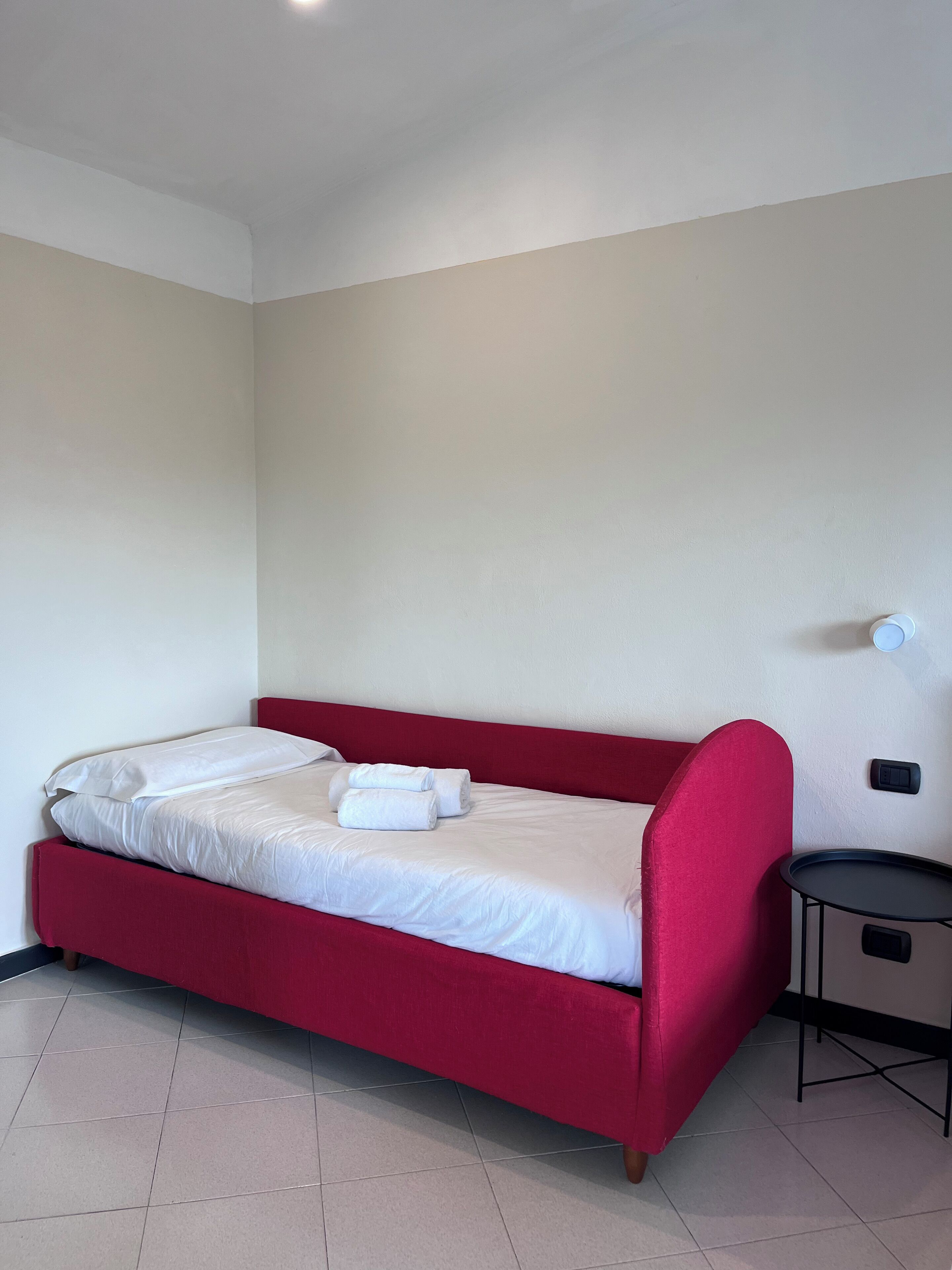 Una cama individual con sábanas blancas y cabecera roja, una mesita redonda pequeña y una lámpara de pared.