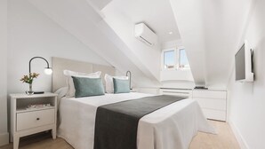 A modern bedroom with a sloped ceiling, a bed with a dark throw, a bedside table with a lamp, and a TV mounted on the wall.