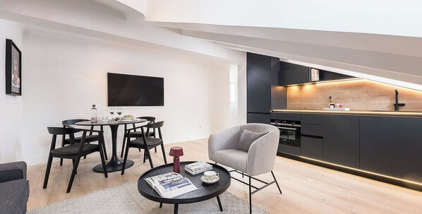 A modern living space with a dining area, a grey armchair, a black coffee table, and a kitchen with a wooden countertop and black cabinets. - Lagasca XLVI - Two Bedroom Apartment, Sleeps 4 (Madrid)