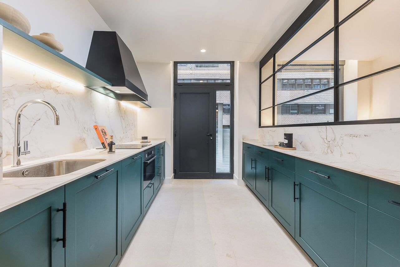 Een moderne keuken met blauwgroene kastjes, een marmeren aanrechtblad en een zwarte deur.