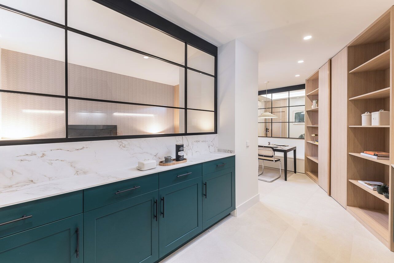 Een moderne keuken met blauwgroene kastjes, een marmeren aanrechtblad en een groot raam met zonwering.