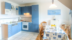 A kitchen with blue cabinets, a dining table with a nautical tablecloth, and a view of the sea through a window. - Fokuhl Apartment with Shared Garden, Balcony, and Wi-Fi (Hooksiel)