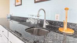 A kitchen with a granite countertop, stainless steel sink, and a wooden utensil holder. There are framed posters on the wall and a small cabinet with a tissue box. - Colorado Riverfront Oasis – Fishing, Views & Perfect Family Getaway (Matagorda)
