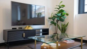 Moderni olohuone, jossa on litteänäyttöinen televisio, tumma puulattia, lasinen sohvapöytä ja ruukussa oleva huonekasvi.