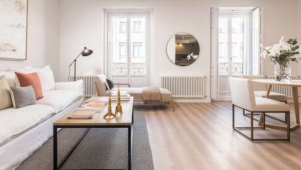 Una sala moderna con un sofá blanco, una mesa de centro, un espejo redondo y un comedor con una silla y una mesa.