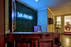 Ett välkomstområde med en neonskylt som visar texten ”SAN ANTONIO”, en receptionsdisk i trä, en inramad flagga och en krukväxt.
