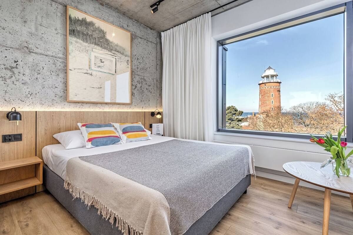 A modern bedroom with a large bed, a view of a lighthouse, and a small table with flowers.