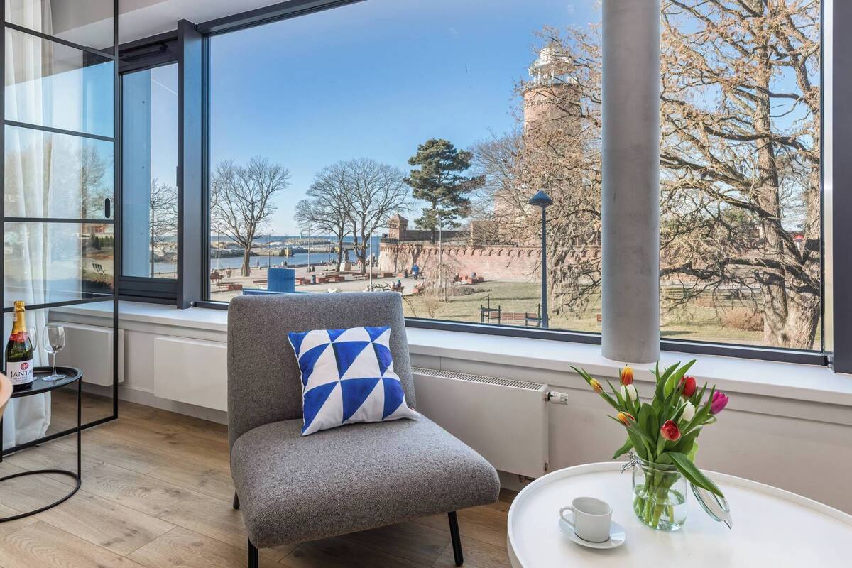 A modern room with a large window, a grey armchair with a blue and white pillow, a small table with a cup and tulips, and a bottle of JANTA wine.