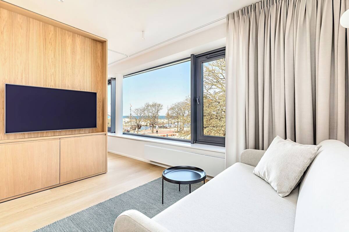 A modern hotel room with a large window, a flat-screen TV mounted on a wooden panel, a sofa with a pillow, and a small round table.