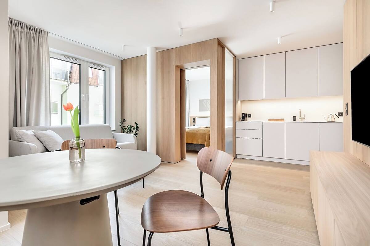 A modern, well-lit living room with a round dining table, a comfortable sofa, and a kitchen area with white cabinets and a flat-screen TV.