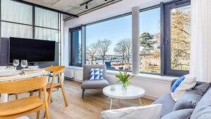 A modern living room with a large window, a flat-screen TV, a dining area with a table and chairs, and a sofa with cushions. - BalticON Apartments Morska (Kolobrzeg)