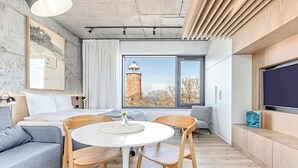A modern, compact living space with a bed, a sofa, a dining table with chairs, a large window with a view of a tower, and a flat-screen TV. - BalticON Apartments Morska (Kolobrzeg)