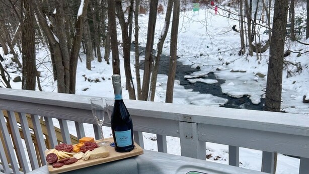 Tinaco con una bandeja de madera que sostiene una botella y aperitivos, rodeado por un paisaje nevado y árboles.