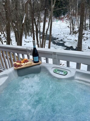 Ein Whirlpool mit einem hölzernen Tablett, auf dem sich eine Flasche und Snacks befinden, umgeben von verschneiter Landschaft und Bäumen.