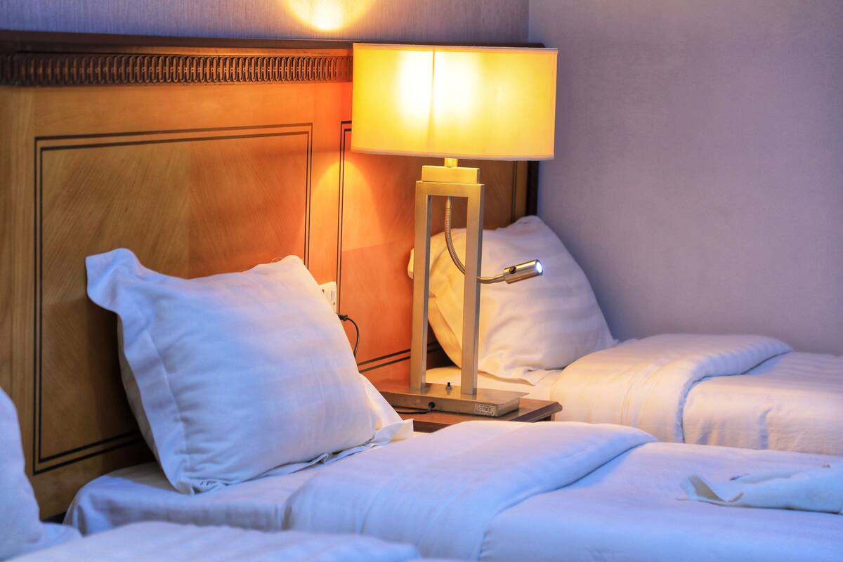 A hotel room with two beds, a wooden headboard, a bedside table with a lamp, and white bedding.