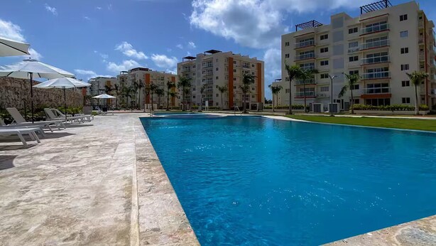 Ein Swimmingpool mit Liegestühlen und Sonnenschirmen, umgeben von mehrstöckigen Gebäuden mit Balkonen und Palmen.
