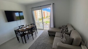 Ein modernes Wohnzimmer mit flachbildfernseher an der Wand, einem Esstisch mit Stühlen und einem grauen Sofa mit gemusterten Kissen.