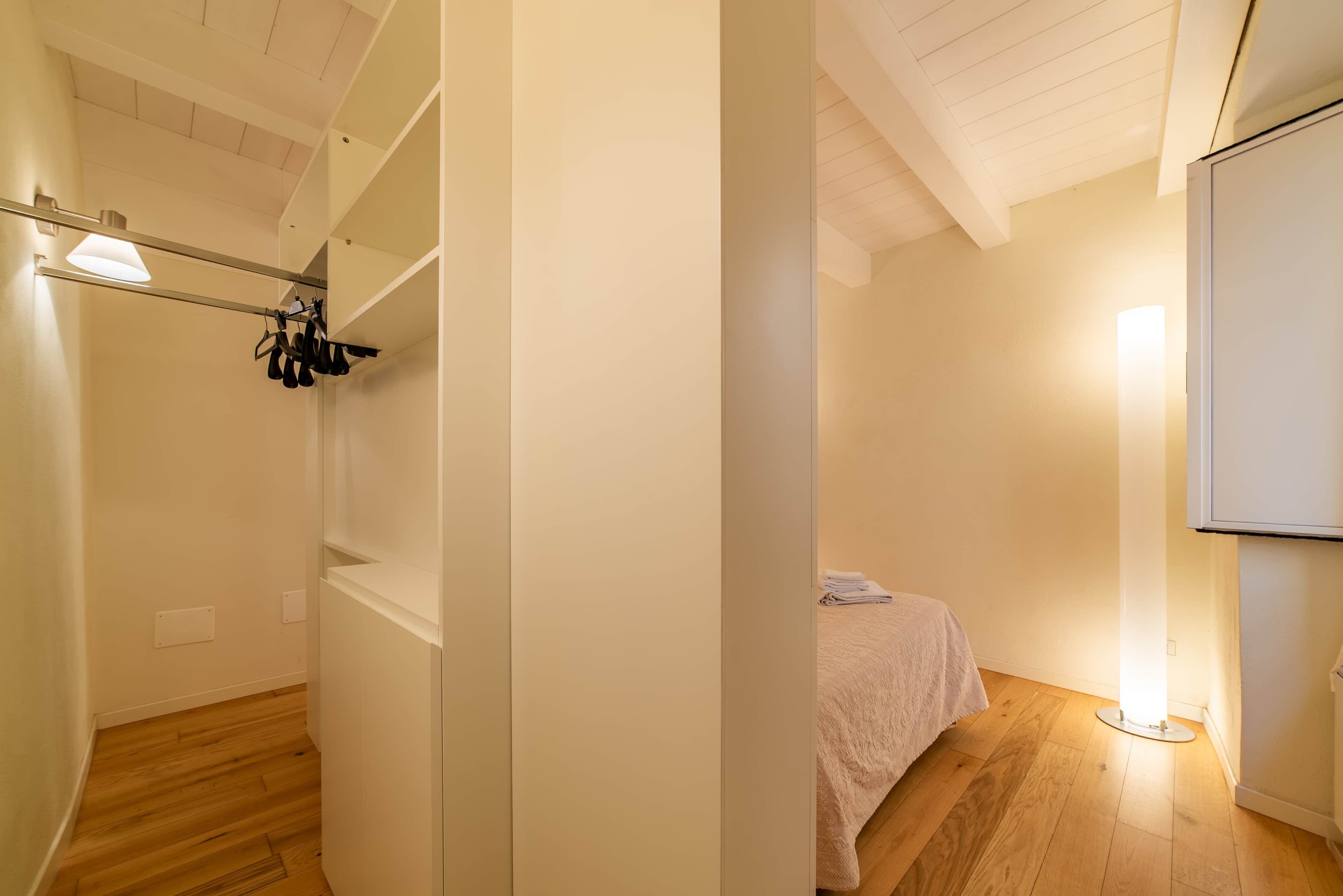 A bedroom with a bed, a bedside table, a nightstand, a wall-mounted shelf, and a ceiling light fixture.