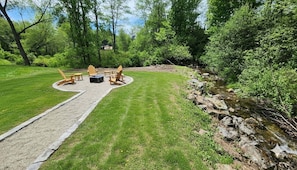 Eine Terrasse mit Adirondack-Stühlen, einem Feuerkorb und einem Steinweg, der zu einem Bach führt.