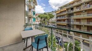 Un balcón con mesa y sillas, con vista a una calle con coches aparcados y edificios de varios pisos.