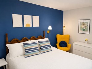 A bed with two pillows, a wooden headboard, a yellow armchair, a bedside table, and a wall with three framed artworks.