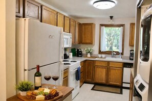 Cocina compacta con armarios de madera, un refrigerador, un microondas, una estufa y un lavamanos. Hay una pequeña ventana con vista a árboles.